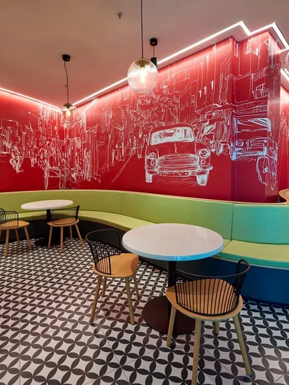 A snippet from a massive 2.5 lac sq ft cafeteria designed to feed 11,000 people daily. This section features a long, curved green banquette and a bold red wall with white line-art graphics of city life.