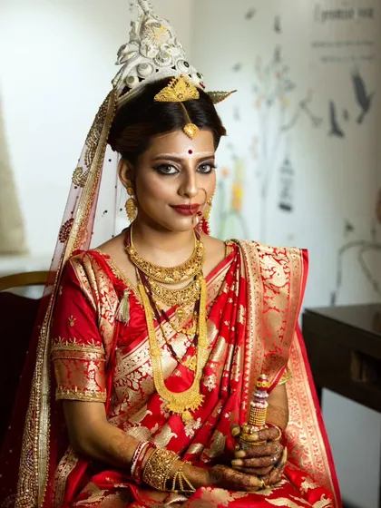 A seated portrait of a Bengali bride holding a 'gach kouto', showcasing her complete traditional attire and jewelry.