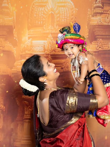 A joyful mother lifts her baby, dressed as Krishna, into the air against a grand temple backdrop.