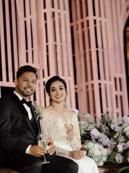 A modern and chic reception stage. The backdrop features a unique pink slatted structure with elegant hanging lights, complemented by a bed of soft purple and white flowers.