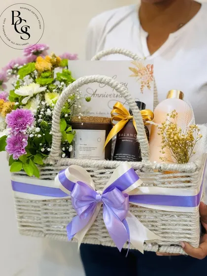 An elegant anniversary hamper in a white woven basket. It's filled with fresh purple and white flowers, scented candles, and bath products, creating a relaxing and romantic gift for a special couple.