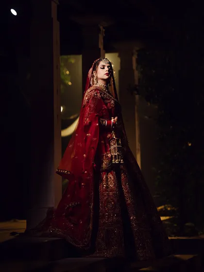 A dramatic and artistic bridal portrait using creative lighting. The bride, in her stunning red wedding lehenga, is captured in a low-light setting, creating a powerful and regal image that feels both timeless and modern.