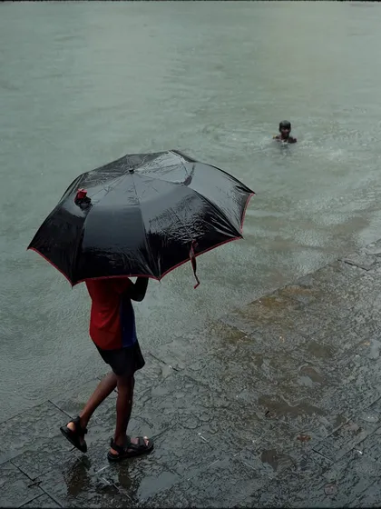 A child with an umbrella walks along the edge of the tank while another swims in the water. The frame tells a story of two different yet simultaneous monsoon activities.