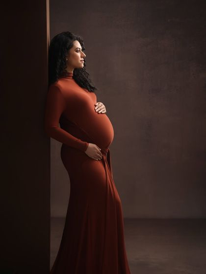A moody and artistic portrait. The deep, earthy red of the gown and the dramatic side lighting create a powerful and contemplative image of motherhood.