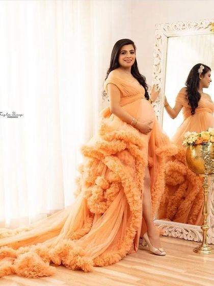 A client posing in a beautiful studio setting with our peach ruffled gown. The off-the-shoulder design and long, textured train make for a very glamorous maternity photo.
