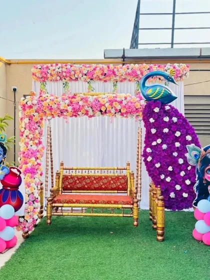An outdoor baby shower setup on a terrace with a beautiful swing (jhula). The decor features a floral arch, Radha Krishna cutouts, and a stunning peacock figure made from artificial flowers.