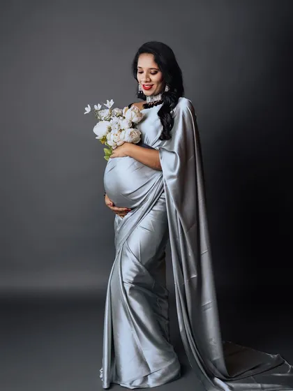 A full-length studio shot of an expectant mother in a graceful silver saree, holding a bouquet of white peonies. The long, flowing drape of the saree adds a touch of regal elegance to the portrait.