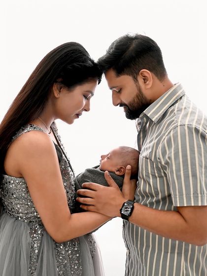 An intimate and artistic newborn family portrait. The parents' heads are touching as they gaze down at their baby, creating a beautiful composition in a high-key white setting.