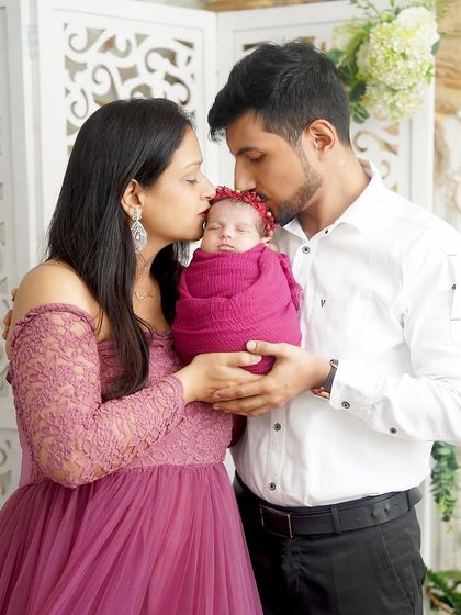 A beautiful family portrait celebrating their newborn daughter. The parents shower their baby with kisses, all dressed in coordinating mauve and pink tones.