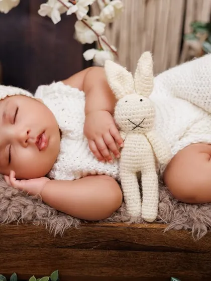 A sleeping newborn in a white knit outfit, cuddling a crochet bunny on a miniature wooden bed. A classic and timeless portrait.