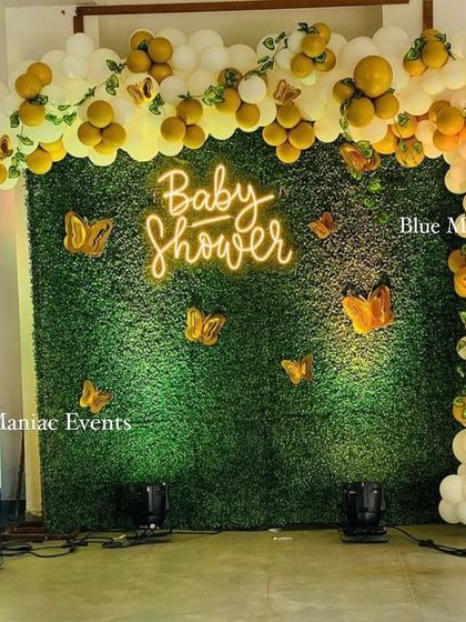 A beautiful baby shower decoration with a green wall backdrop. The arch of white and yellow balloons is adorned with butterfly cutouts, and a 'Baby Shower' neon sign adds a warm glow.