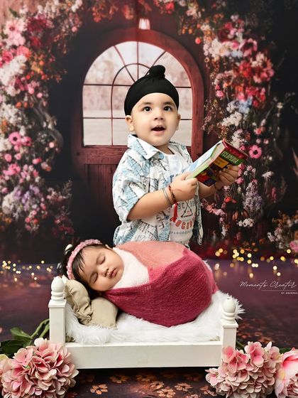 A storybook moment between siblings. An older brother reads a book to his newborn sister as she sleeps peacefully in a tiny bed, surrounded by a magical floral arch.