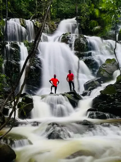 A long exposure shot captures two trekkers standing like statues amidst the silky flow of a beautiful waterfall.