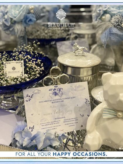 A close-up of the personalized announcement card for a baby boy. The hamper includes a modern white teddy bear with a bow tie, a silver jar with a rocking horse lid, and other elegant blue and silver details.