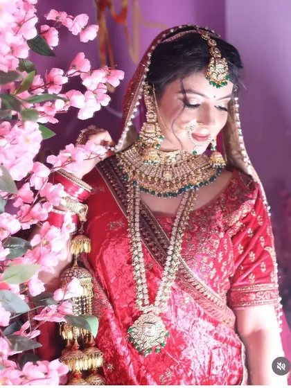 A bride surrounded by pink flowers, wearing a traditional red lehenga with heavy green and gold jewellery. The combination of colors creates a classic and beautiful bridal look.