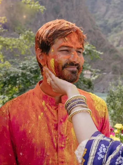 A bride's hand caresses the groom's face, both covered in holi colors, with the Ganges in the background. This colorful, intimate moment captures the spirit of their wedding in Rishikesh.