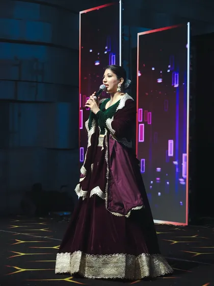 Dressed in a royal purple and green velvet lehenga, I am addressing the audience. My attire is always chosen to complement the theme and grandeur of the event.