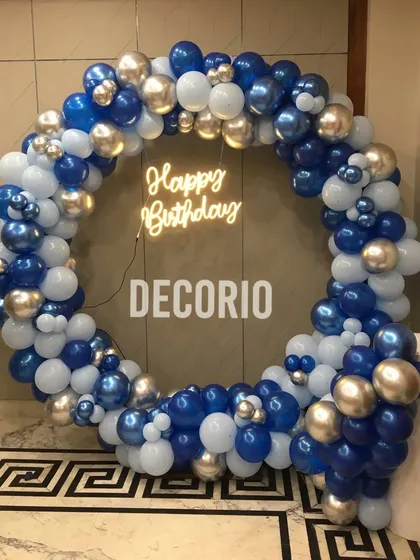 A full circular balloon arch in shades of royal blue, light blue, and silver. A 'Happy Birthday' neon sign in the center makes this a perfect, eye-catching photo backdrop.