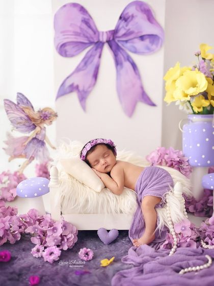 A whimsical, fairy-themed newborn setup in shades of purple. The sleeping baby is surrounded by magical elements like fairies, mushrooms, and a giant bow.