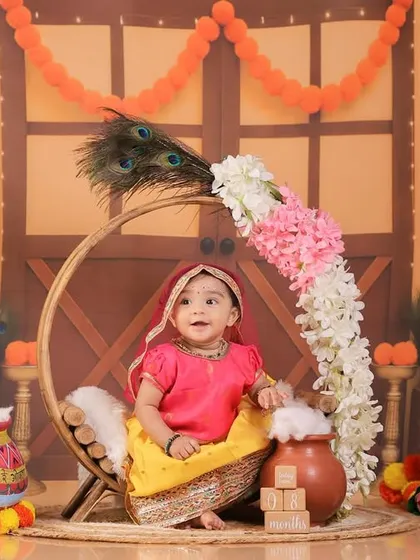 The little girl from the Radha-themed shoot, sitting in a decorated circular swing, surrounded by marigold flowers and peacock feathers.