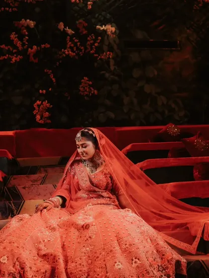 A stunning portrait of the bride, Sakshi, seated in her magnificent red lehenga. The dramatic lighting and rich colors make her look like royalty.