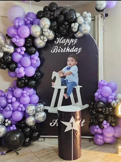 A stylish first birthday party with a black, purple, and silver color scheme. This setup includes a personalized photo backdrop, a rocking horse prop, and a light-up number '1'.