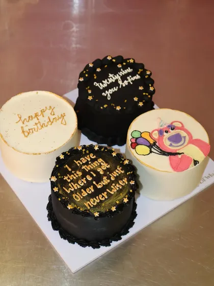A chic and modern set of four bento cakes in a black, white, and gold color scheme, featuring fun quotes and a cute Lotso bear illustration.