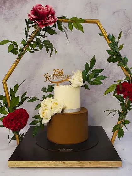 A sophisticated two-tier cake for a 25th silver jubilee anniversary, presented within a decorative golden hexagonal frame. The clean white top tier contrasts beautifully with the smooth brown bottom tier, adorned with fresh white roses.
