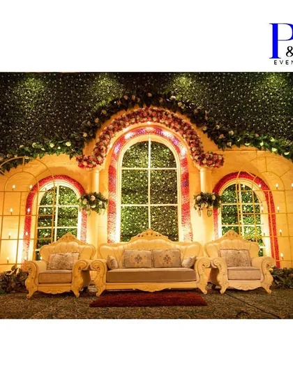 A grand reception stage setup with a backdrop of lush green foliage and floral arches. The elegant sofa seating and warm lighting create a sophisticated and inviting space for the couple.