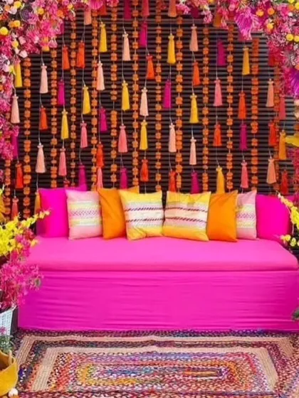 A vibrant and colorful Mehandi seating area with a pink diwan and a floral arch. The setup includes a traditional woven rug and baskets of fresh flowers.
