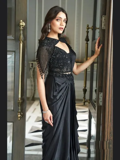 For a chic and modern reception or cocktail look, this black pre-draped saree is a perfect choice. It combines the elegance of a saree with the ease of a gown, featuring a stunningly embellished blouse with a stylish cutout and cape sleeves.