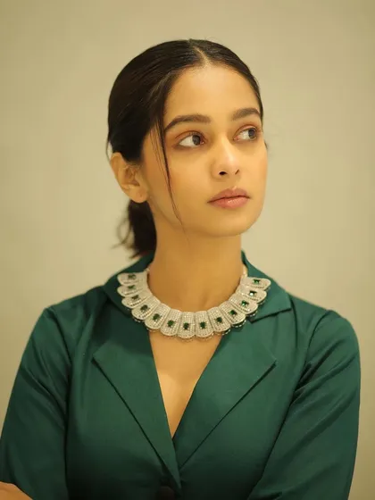 A close-up portrait of Mugdha Chaphekar, highlighting the clean makeup and elegant low ponytail. The focus is on her expressive eyes and the statement necklace.