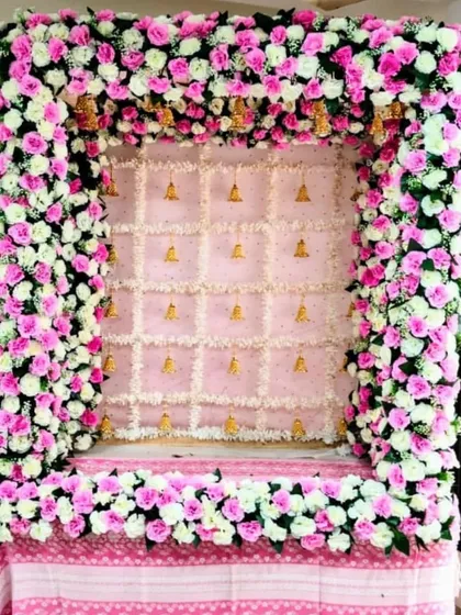 A beautiful pink and white floral frame designed for a home Ganpati setup. The backdrop is made of delicate white flowers with small golden bells, perfectly complementing the dense rose arrangement.
