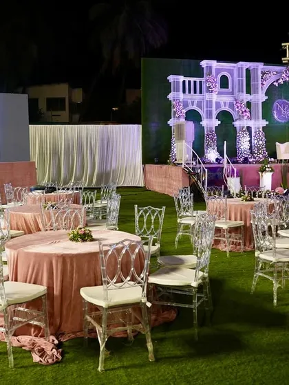 A wide view of the wedding reception setup, showing the guest seating arrangement with clear acrylic chairs and blush pink linens. In the background, you can see the grand stage and a large LED screen for displaying photos and videos.