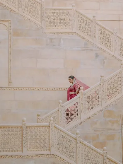 A bride's graceful descent. Dressed in a beautiful pink lehenga, she walks down the stone staircase of the palace, a timeless and elegant portrait moment before her ceremony.