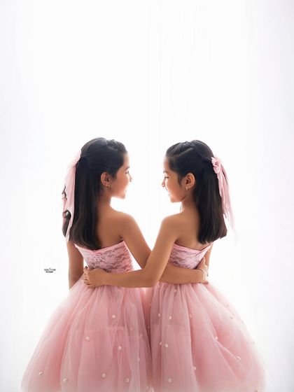 A quiet, connected moment between two sisters in their beautiful pink tulle dresses. A simple, high-key background keeps the focus entirely on them.