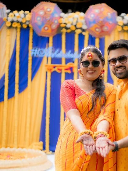 Akshat and Divyanshi pose with their henna-adorned hands, their matching yellow and orange outfits perfectly complementing the Haldi decor.
