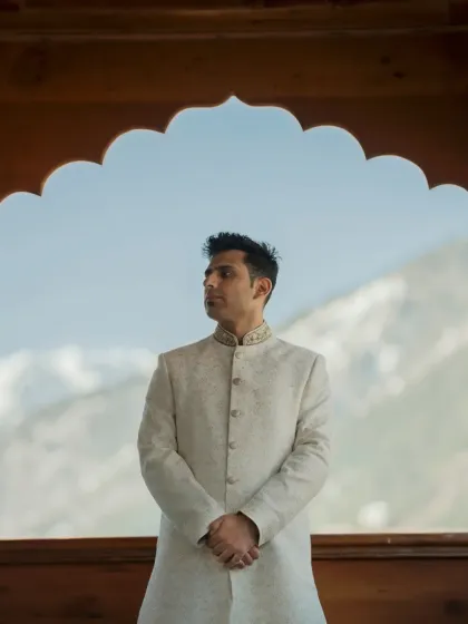 A handsome portrait of the groom in his ivory sherwani, framed by a traditional wooden archway against the mountain view.