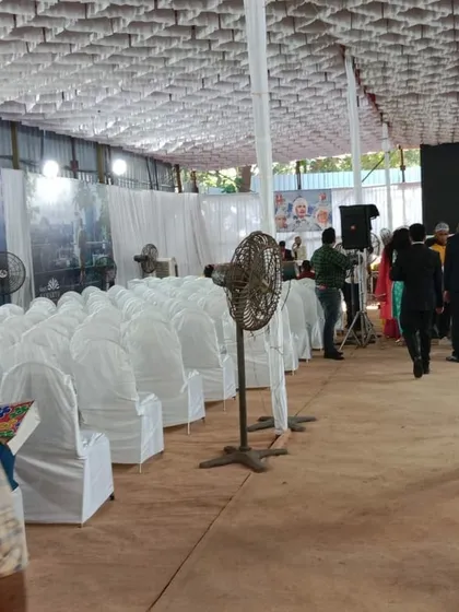 This image shows a wide view of a corporate event in progress. You can see the neat rows of fully covered chairs, pedestal fans for cooling, and fabric draped walls that create a professional environment inside a basic hall.