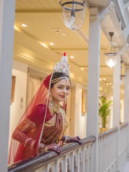 A beautiful portrait of a Bengali bride in her traditional attire and mukut, looking out from a balcony with a gentle smile.