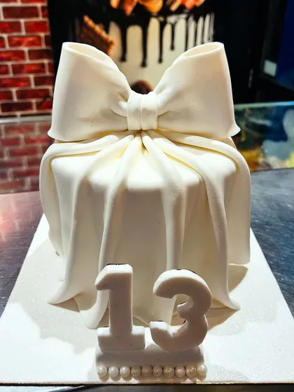 An elegant all-white cake for a 13th birthday, designed to look like a gift box with a large, flowing bow.