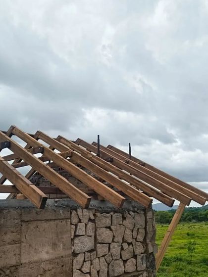 A building takes shape in harmony with its surroundings. Here, walls made of natural stone and rammed earth rise from the ground, while the wooden framework for the roof is put in place against a cloudy sky.
