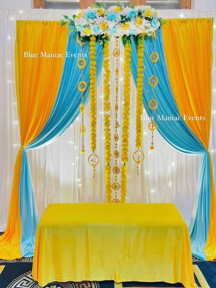 A bright and cheerful Haldi setup using yellow and sky blue drapes. The marigold garlands and white floral border add a classic touch to this fresh color combination.