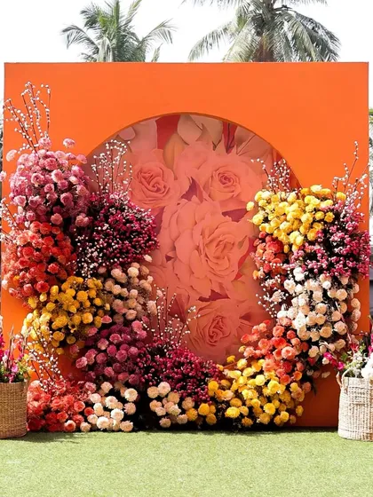 A beautiful floral photo booth, with a wreath of colorful flowers framing a translucent center, set against a bright orange panel.
