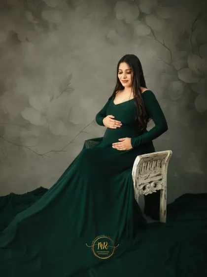 A graceful seated pose in a deep green maternity gown. The long train drapes elegantly around a rustic white chair, creating a timeless and sophisticated pregnancy portrait.
