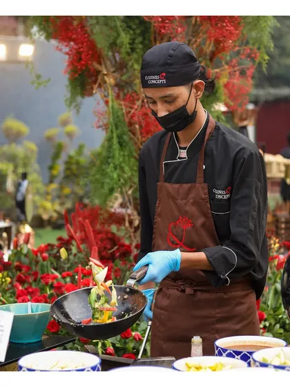 A chef tosses fresh vegetables in a wok at an outdoor live station surrounded by beautiful red floral decor. This highlights our ability to create a complete sensory experience where the food preparation complements the event's theme.