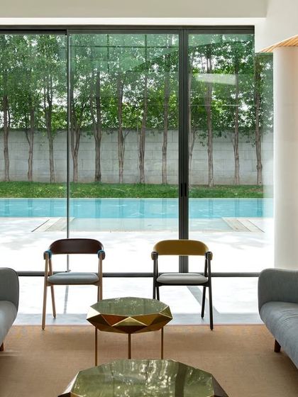 A minimalist living room with a floor-to-ceiling glass wall that frames a view of the swimming pool and a line of trees. The design creates a powerful connection to the outdoors, making the landscape an integral part of the interior.