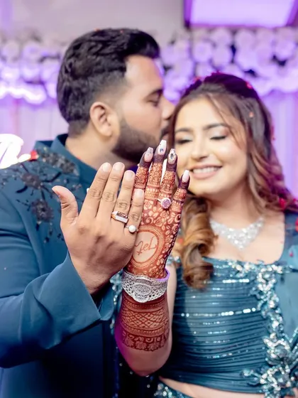 A happy couple at their engagement. The groom kisses his bride's forehead as they both show off her beautiful back-hand mehendi with their initials.