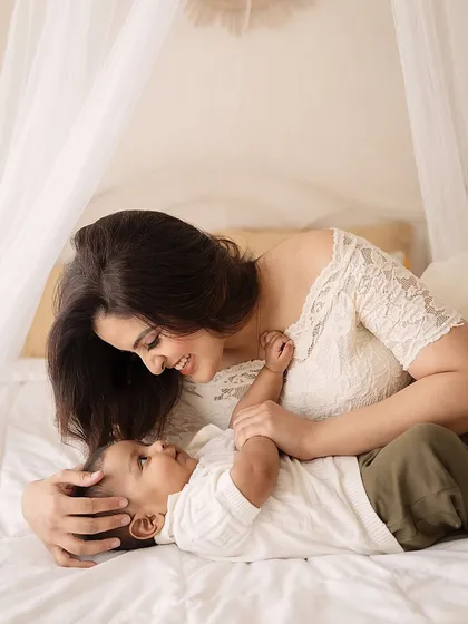 A mother and her baby share a quiet, loving gaze on a soft, white bed. The delicate lace and sheer canopy add to the dreamy, ethereal feel of this portrait.