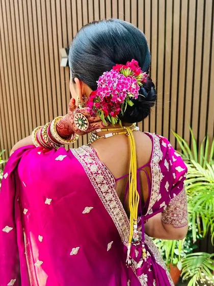 A classic bridal bun elevated with a vibrant pop of fresh pink flowers. This hairstyle is perfect for complementing a traditional Indian wedding lehenga or saree.